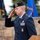 A U.S. Air Force officer in dress blues is rendering a salute at what appears to be an outdoor military ceremony. He is standing at a podium and wearing a black beret with a unit flash patch, along with multiple service ribbons and badges. Blue military flags are visible in the blurred background.