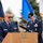 A U.S. Air Force officer, identified by nameplate as 'MEGAS', is speaking at a podium during an outdoor military ceremony. He is wearing the Air Force Service Dress uniform with a chaplain cross insignia and multiple service ribbons. A second uniformed military member stands in the background near color guard flags.