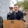 A man in a dark navy suit with a burgundy tie is speaking at a wooden podium equipped with multiple microphones. The setting appears to be an outdoor military or veterans memorial ceremony, with a uniformed military officer visible in the background. Bronze statues and stone memorial walls with plaques are visible in the background.