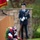 A memorial ceremony is depicted with a patriotic wreath in red, white, and blue flowers prominently displayed in the foreground on a stand. In the background, a uniformed military service member stands at attention near a stone wall and podium. The setting appears to be an outdoor memorial or commemorative event, likely associated with a U.S. military observance such as Memorial Day or Veterans Day.