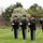 Four U.S. Army soldiers in dress uniforms are standing at attention on a manicured green lawn during what appears to be a formal military ceremony. Three soldiers are holding rifles at order arms position while the fourth, on the right, is rendering a salute. The setting includes a brick building, flagpole, and blooming spring trees in the background.
