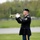A U.S. Army soldier in dress uniform stands on a grass field playing a brass bugle or trumpet. The soldier appears to be performing a ceremonial duty, likely playing 'Taps' at a military memorial or funeral service. The setting is an open outdoor area with trees visible in the background.