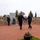 A military memorial ceremony is taking place at an outdoor veterans memorial site. Two individuals — one in military dress uniform and one in civilian attire — are carrying a red, white, and blue wreath toward a monument, while a uniformed officer stands at salute to the left. The memorial features a stone relief wall, a kneeling soldier sculpture, commemorative plaques, and multiple flags.