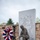 A military memorial monument featuring a bronze kneeling soldier statue in front of a granite stele engraved with soldiers from different eras of American military history. A 'Battlefield Cross' (boots, rifle, and helmet) is displayed on sandbags on the monument's base, and a red, white, and blue floral wreath on a stand is placed to the left. Bronze memorial plaques are mounted on a stone wall in the background.