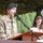 A U.S. Army officer in OCP (Operational Camouflage Pattern) uniform is speaking at a podium with a microphone during what appears to be an official military ceremony or event. He is wearing a black beret and has a 101st Airborne Division patch visible on his left shoulder along with a U.S. flag patch. A civilian woman in a white top stands behind him, appearing to hold papers, possibly serving as an interpreter or co-presenter.