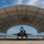 A military fighter jet, identifiable as an F-35 Lightning II, is positioned under a large arched fabric hangar shelter on a flight line. Ground crew personnel are visible around and on top of the aircraft, performing what appears to be pre-flight or post-flight maintenance. The image is taken from a low, wide-angle perspective that emphasizes the symmetrical arch of the shelter structure.