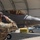 A military ground crew member in MultiCam/OCP camouflage uniform is saluting an F-35 Lightning II fighter jet (tail number 5078) as it prepares to taxi out of a hardened aircraft shelter or portable hangar. A pilot is visible in the cockpit. The scene appears to be at a U.S. Air Force base on a clear, sunny day.