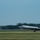 An F-35 Lightning II fighter jet is captured in motion on a runway, likely during takeoff or landing. The aircraft bears 'LF' tail codes, indicating assignment to Luke Air Force Base in Arizona. Motion blur in the background emphasizes the aircraft's speed.