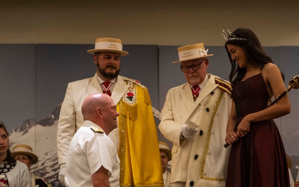 A formal ceremonial event is taking place on a stage, likely a pageant or civic coronation ceremony. A person in a white military-style uniform is kneeling before a young woman wearing a tiara and holding a scepter, who stands on a platform. Two officials in white suits with decorative regalia and straw boater hats oversee the proceedings, while attendants seated in the background hold floral bouquets.