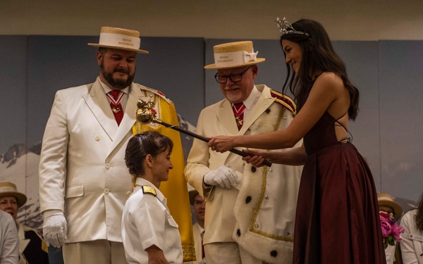 A formal ceremonial event is taking place on a stage, where a person in a white naval/military uniform is kneeling while a woman wearing a tiara and dark burgundy gown touches them with a ceremonial sword or scepter. Two men in white ceremonial jackets and straw boater hats bearing insignia are standing nearby as officials. An audience of seated attendees, many in white attire and holding floral bouquets, observes the ceremony in the background.