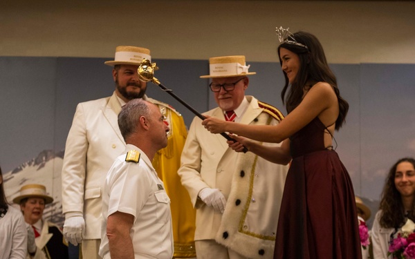 A formal ceremonial event is taking place on a stage, where a woman wearing a tiara and burgundy gown is touching a scepter or sword to the shoulder of a man kneeling before her in a white naval or military uniform. Two men in white suits and straw boater hats stand behind them, one holding a ceremonial staff. Seated attendants in matching white jackets holding pink flowers are visible in the background.