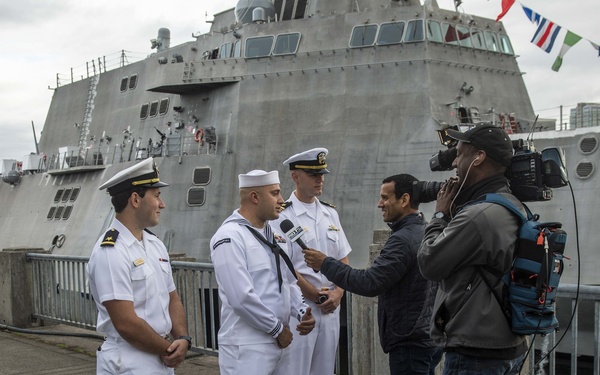 Three U.S. Navy personnel in white dress uniforms are being interviewed by a television news crew on a dock. A large gray naval warship, decorated with signal flags, is visible in the background. The setting appears to be a public port event, possibly a fleet week or ship commissioning ceremony.