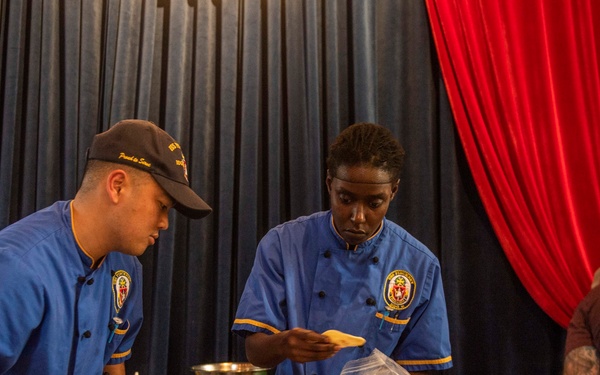 Two individuals wearing matching blue chef uniforms with military-style insignia patches are plating dishes at a culinary competition or event. Multiple white plates with green noodle/salad dishes are lined up on a wooden table in the foreground. A sign visible on the right indicates 'Team 5' with a partial ingredients list, suggesting a structured cooking competition format.