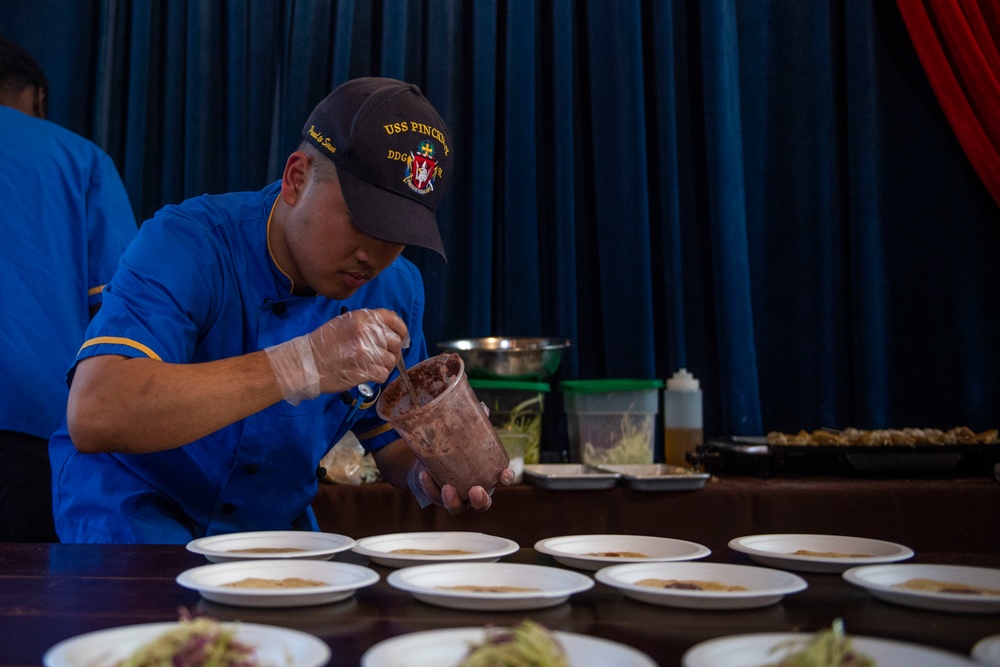 A person wearing a USS Pinckney DDG cap and blue uniform is carefully plating food onto white dishes arranged in a row on a table. The setting appears to be a culinary competition or formal food service event, with multiple plates being prepared simultaneously. The individual is using gloved hands and a cup mold to portion food onto the plates.