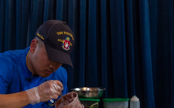 A person wearing a USS Pinckney DDG cap and blue uniform is carefully plating food onto white dishes arranged in a row on a table. The setting appears to be a culinary competition or formal food service event, with multiple plates being prepared simultaneously. The individual is using gloved hands and a cup mold to portion food onto the plates.