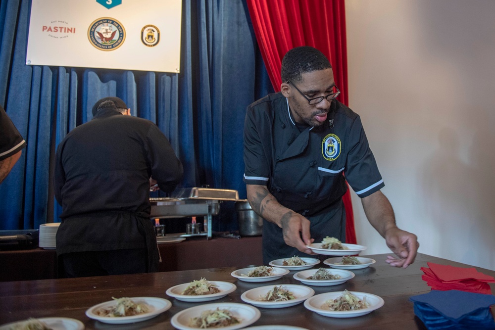 A Navy chef in a black uniform with a military insignia patch is plating multiple dishes on white plates arranged on a wooden table. A second person in black is working in the background near chafing dishes. The setting appears to be a military culinary event or demonstration, indicated by a banner showing 'PASTINI' and U.S. Navy/Department of the Navy seals.