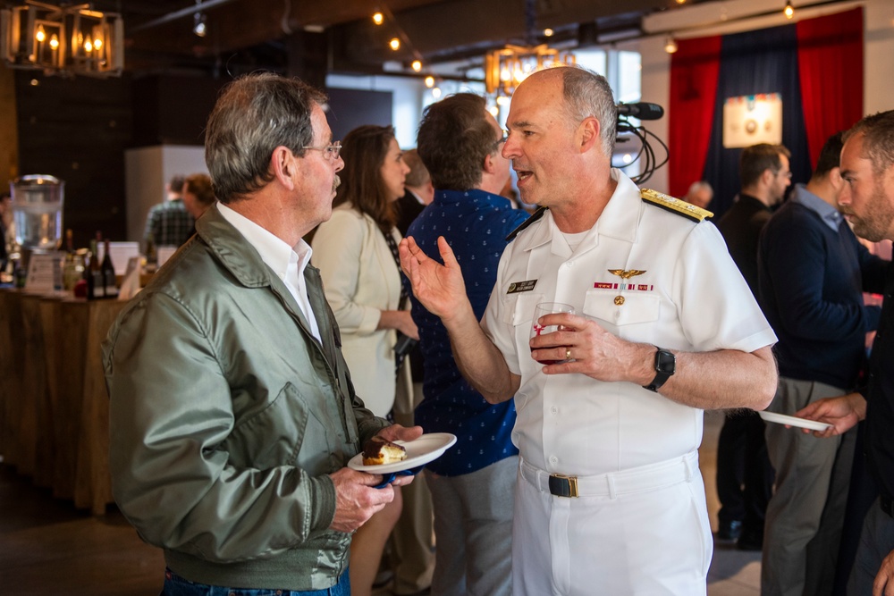 A social gathering or reception event where a U.S. Navy officer in white dress uniform is engaged in conversation with a civilian man wearing a green jacket. The officer displays visible rank insignia, aviator wings, and service ribbons consistent with a senior flag officer. The setting appears to be an indoor venue decorated with string lights and draped fabric.