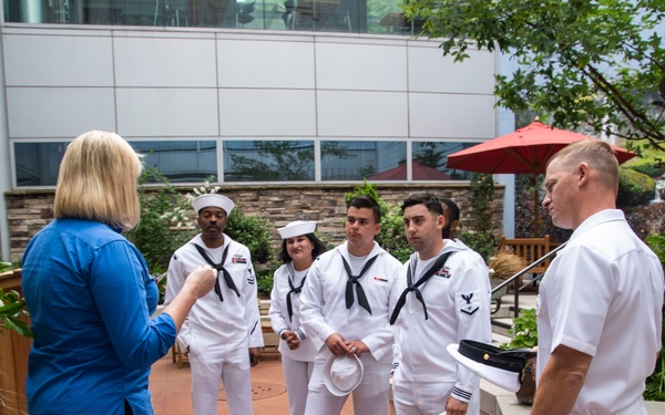 A woman in a blue shirt is speaking to a group of five U.S. Navy personnel dressed in white dress uniforms in an outdoor courtyard setting. The group includes four enlisted sailors and one senior officer, several of whom are holding their white covers (hats) in their hands. The setting appears to be a modern institutional or medical facility with a landscaped garden area.