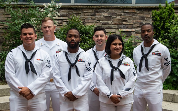 Six U.S. Navy personnel are posed together in a formal group photograph outdoors. They are wearing Service Dress White uniforms with black neckerchiefs, and several individuals display ribbons and rating badges on their uniforms. The group consists of five men and one woman, standing in front of a stone wall with lush green vegetation.