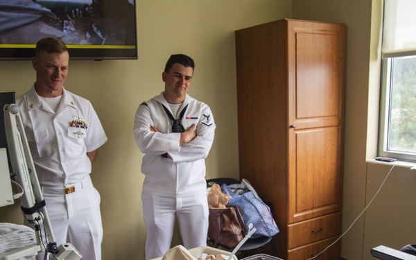 Two U.S. Navy personnel in white dress uniforms are visiting a patient in a hospital room. The patient is lying in a hospital bed with medical equipment nearby, and a bedside table holds various items including a water pitcher, mug, and documents. The setting appears to be a morale or community outreach visit.