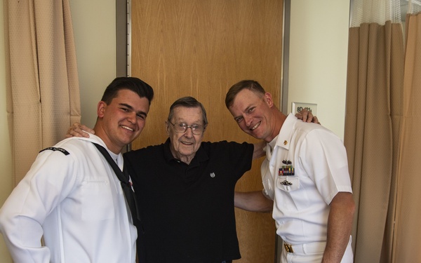 Two U.S. Navy personnel in white dress uniforms are posing with an elderly civilian man in what appears to be a medical or care facility room. The sailor on the left wears enlisted dress whites, while the officer on the right displays multiple ribbons and insignia on his uniform. All three individuals are smiling and appear to be in good spirits during what looks like a visit.