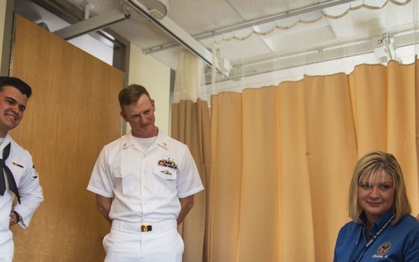 Two U.S. Navy personnel in white dress uniforms are visiting an elderly patient in what appears to be a medical facility or veterans hospital room. A civilian woman wearing a blue polo shirt with a lanyard/badge is seated beside the elderly man, holding paperwork. The group appears to be engaged in a friendly conversation.