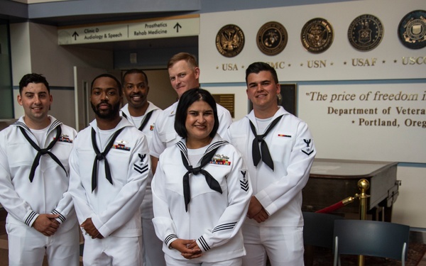 Six U.S. Navy sailors in white dress uniforms (Dress Whites) are posing for a group photo inside the Department of Veterans Affairs facility in Portland, Oregon. They are standing in front of a wall displaying military branch seals (USA, USMC, USN, USAF, USCG, USMM) and the quote 'The price of freedom is visible here.' The group consists of five males and one female, all appearing to hold Petty Officer ranks based on visible chevrons.
