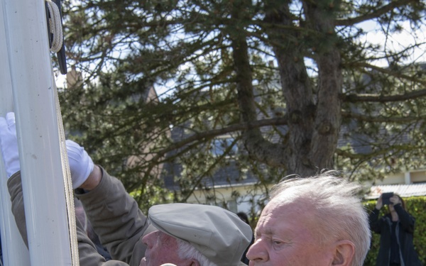 Two elderly men, appearing to be military veterans based on their service medals, are participating in a flag-raising ceremony at an outdoor location. The foreground figure wears a dark suit with multiple medals and white ceremonial gloves, handling a rope on a flagpole. A photographer is visible in the background documenting the event.