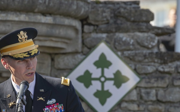A U.S. Army general officer named ZINN is delivering a speech at a formal military ceremony, reading from a document at a microphone. Behind him is a stone wall displaying the insignia of the 4th Infantry Division (ivy leaf patch on a white diamond). The setting appears to be an outdoor memorial or commemorative event, likely in Europe based on the stone architecture.