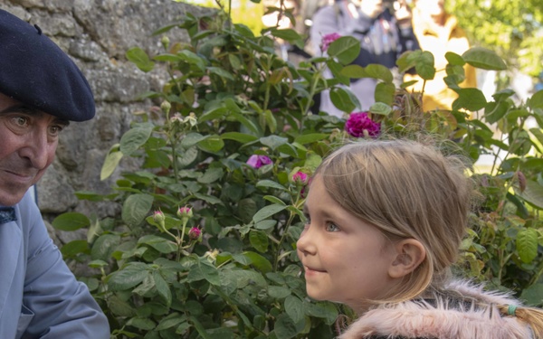 A young blonde girl in a gray coat with pink fur trim stands near a white bucket containing white roses decorated with a red, white, and blue ribbon. She holds a small American flag and appears to be participating in a commemorative or ceremonial event. An elderly man wearing a dark beret and light blue jacket crouches beside her near a stone wall.