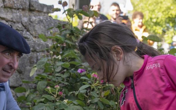 A young girl wearing a pink Kappa jacket leans forward to arrange or place white roses in a white container with what appears to be a tricolor ribbon. An older man wearing a blue beret and gray jacket watches from the left. A crowd of onlookers is visible in the background near a stone wall.