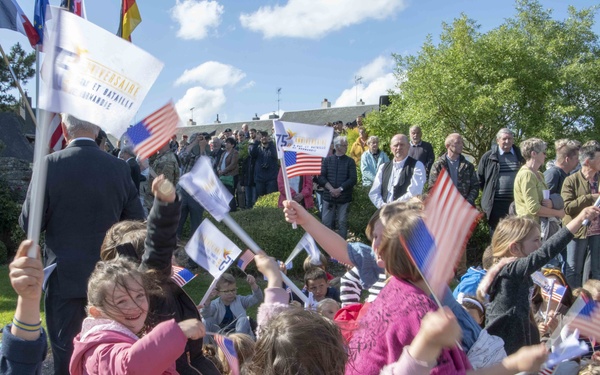 A festive outdoor commemoration event featuring children enthusiastically waving American flags and event banners. The gathering appears to be a D-Day anniversary ceremony in Normandy, France, based on the visible banner text referencing 'Bataille de Normandie' and '75e Anniversaire' (75th Anniversary). A large crowd of adults stands in the background near a stone monument surrounded by greenery.