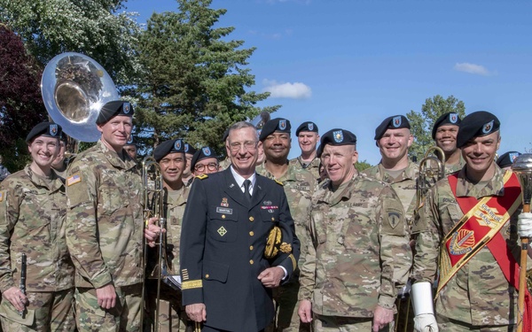 A group of U.S. Army soldiers in camouflage uniforms pose for a group photograph with one senior officer in dress uniform at the center. The soldiers appear to be members of a military band, as several are holding brass instruments including French horns, a trombone, and a sousaphone. The setting is an outdoor garden or park area with green grass and trees in the background.