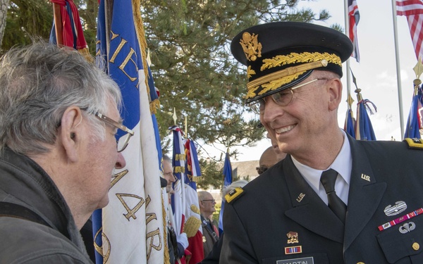 A U.S. Army officer in dress uniform, identified by his name tag as 'MARTIN', is shaking hands and smiling with a civilian man at what appears to be a formal military ceremony or commemoration event. The setting includes multiple national and military flags in the background, suggesting an official outdoor ceremony, possibly at a memorial site. The officer wears senior officer insignia including a general officer's cap with gold braid.