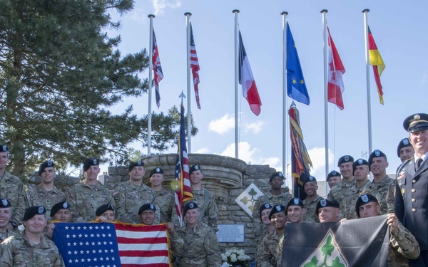 A group of U.S. Army soldiers pose for a formal group photograph at a memorial monument, holding a 48-star American flag and a 4th Infantry Division banner. The setting appears to be a commemorative site, likely in Normandy, France, given the multiple national flags visible in the background. Floral wreaths are placed at the base of the stone monument, suggesting a memorial ceremony has taken place.