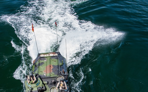 An aerial top-down photograph of a military amphibious assault vehicle (AAV) traveling at speed across open dark green water. The vehicle displays green camouflage paint and has multiple personnel visible on its deck, along with two antenna masts, one bearing a small red and yellow flag. A prominent white wake trails behind the vessel indicating significant forward speed.