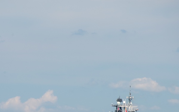 A large gray military amphibious assault ship is anchored or underway on open blue water under a partly cloudy sky. The vessel features a flat flight deck with aircraft visible on top, a prominent island superstructure with masts and communications equipment, and an open stern well deck. A smaller vessel is visible in the distant background on the horizon.