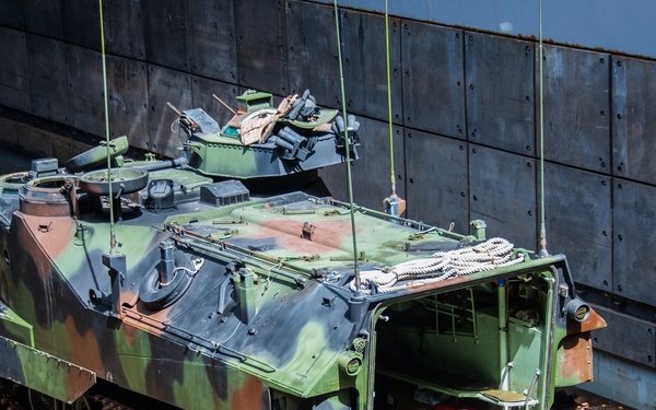 An amphibious assault vehicle (AAV), likely an AAV-7/AAVP7A1, is positioned on what appears to be the deck of a naval vessel or landing ship. The vehicle is painted in a three-color camouflage pattern (green, brown, black) and has its rear ramp lowered, revealing the interior troop compartment. Lifting cables are attached to the vehicle, suggesting it is being loaded or offloaded.