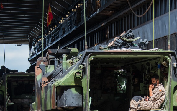 A military personnel member is seated inside the open rear ramp of a tracked armored personnel carrier (APC), likely an AAV7 or similar amphibious assault vehicle, painted in green camouflage. The vehicle is positioned inside what appears to be the well deck of a naval amphibious assault ship. A second similar vehicle is visible in the background.