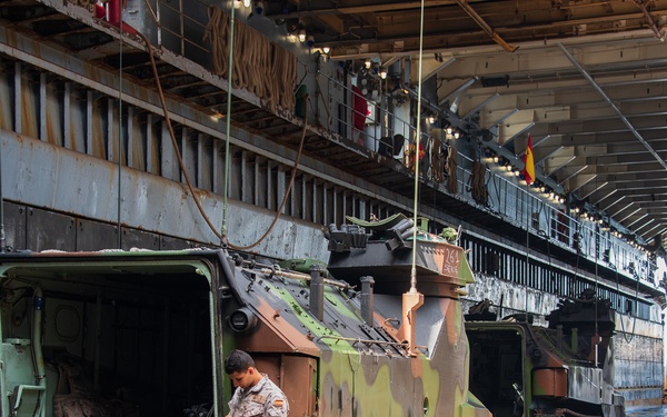 A military personnel member is stepping off or inspecting the rear ramp of a camouflaged tracked armored vehicle, likely an M113 or similar APC variant, inside the well deck of a naval amphibious vessel. The vehicle displays a green, brown, and tan camouflage pattern with a Spanish flag patch visible on the soldier's uniform. At least one additional armored vehicle is visible in the background within the same well deck.