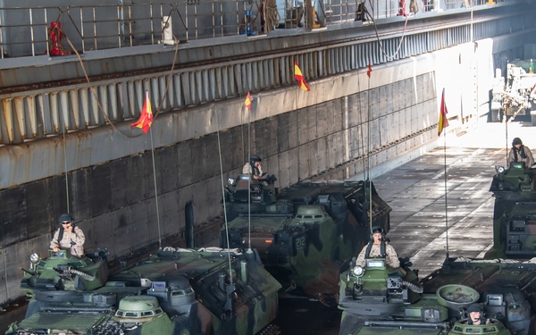 The image shows multiple amphibious assault vehicles (AAVs) staged inside what appears to be the well deck of a naval amphibious transport ship. The vehicles are in green and brown camouflage paint with visible unit numbers (112, 122, 212) and are crewed by military personnel visible in the hatches. Red and yellow flags are positioned along the deck, likely serving as lane markers or guidance signals.
