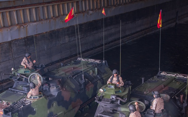 Three amphibious assault vehicles (AAVs) are staged inside what appears to be the well deck of a naval amphibious ship, preparing for a water launch operation. Each vehicle is crewed by military personnel in desert camouflage uniforms and helmets, with red and yellow pennant flags mounted on antenna masts. The vehicles bear visible unit numbers (112 and 122) and are positioned on a flooded or partially flooded deck platform.