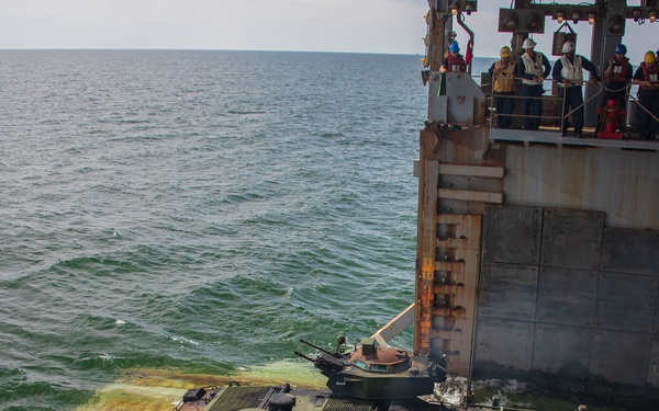 An amphibious armored vehicle numbered '212' with green camouflage paint is exiting the stern well deck of a naval vessel into open water. A Spanish flag is visible on the ship's hull, indicating this is a Spanish Navy operation. Personnel wearing safety gear including helmets and life vests observe from the upper deck of the ship.