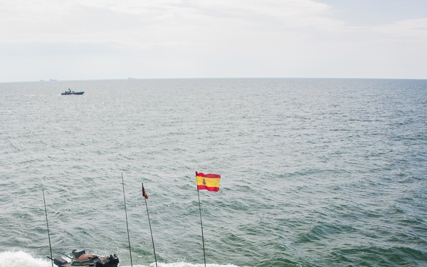 A Spanish military amphibious armored vehicle is launching from a naval vessel's ramp into open sea, creating significant white water spray. The vehicle displays the Spanish flag (red and yellow) and appears to be conducting an amphibious assault exercise or military operation. A naval vessel is visible in the background on the horizon.