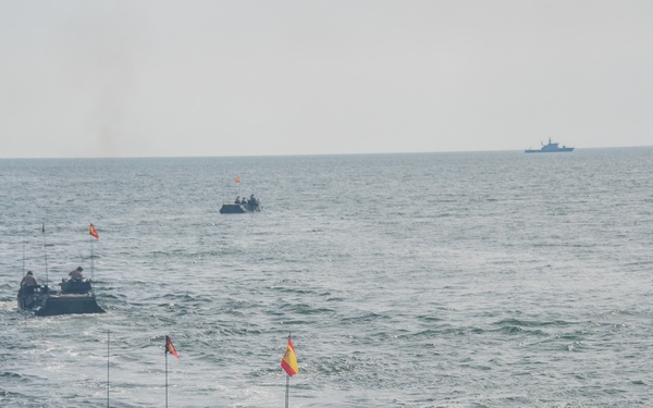 The image shows a formation of amphibious assault vehicles (AAVs) traversing open sea water, each flying what appears to be the Spanish flag (red and yellow horizontal stripes). At least four vehicles are visible in the foreground and middle distance, with a larger naval vessel visible on the horizon in the upper right. This appears to be a Spanish Marine Corps amphibious military exercise or operation.