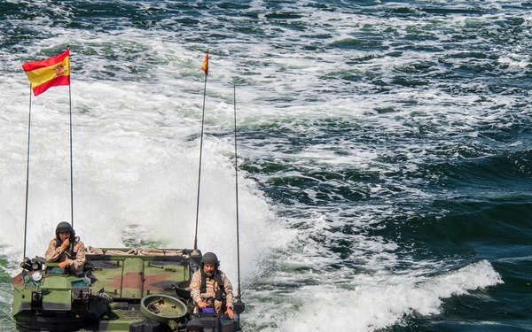 Two amphibious assault vehicles (AAVs) are traversing open water at speed, displaying Spanish national flags. Military personnel in combat uniforms and helmets are visible operating the vehicles. The image appears to document a Spanish military amphibious exercise or operation.