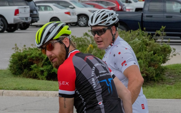 Two male cyclists are stopped at what appears to be a crosswalk in a parking lot area. The foreground cyclist wears a yellow-green Giro helmet, red and black jersey with 'Team Tyler Hamilton' branding and 'Warrior Rides' text, while the background cyclist wears a white helmet and white jersey. Both are equipped with cycling sunglasses and appear to be mid-conversation or pausing during a ride.