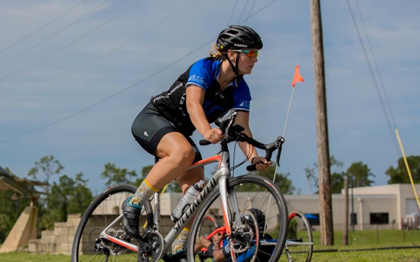 A female cyclist rides a Specialized road bike on a paved road, wearing a blue and black cycling kit, black helmet, and sunglasses. Behind her, a second cyclist on a recumbent or hand-cycle style bike is visible. The setting appears to be a cycling event or race in a semi-rural area with utility poles, grass, and buildings in the background.