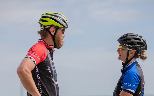 Two cyclists in professional cycling attire are engaged in conversation outdoors near a flat, open area. The male cyclist on the left wears a red and black jersey with 'Warrior Games' and 'Department of Defense' text visible, while the female cyclist on the right holds a Specialized road bike. Both wear helmets and sunglasses, suggesting they are either before or after a cycling event.