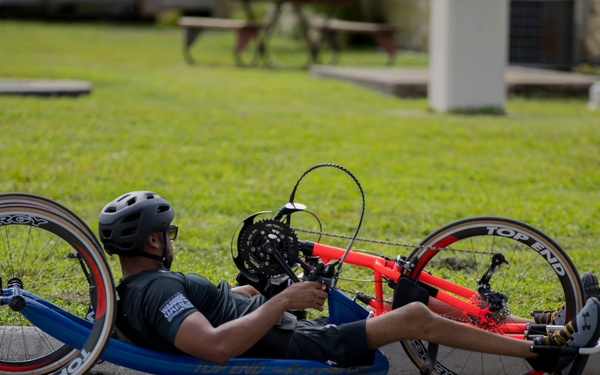 A person is riding a recumbent hand cycle on a paved road, lying in a reclined position and propelling the vehicle using hand cranks. The hand cycle is a 'Top End' brand racing model with red frame and Spinergy wheels. The rider is wearing a black helmet and dark athletic clothing.